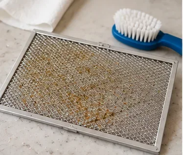 A removed microwave grease filter on a countertop showing visible brown grease buildup in the metal mesh.