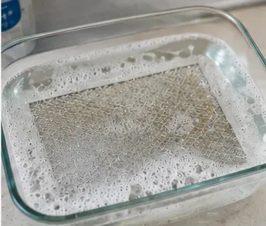 A metal microwave grease filter soaking flat in a glass pan of hot, soapy water.