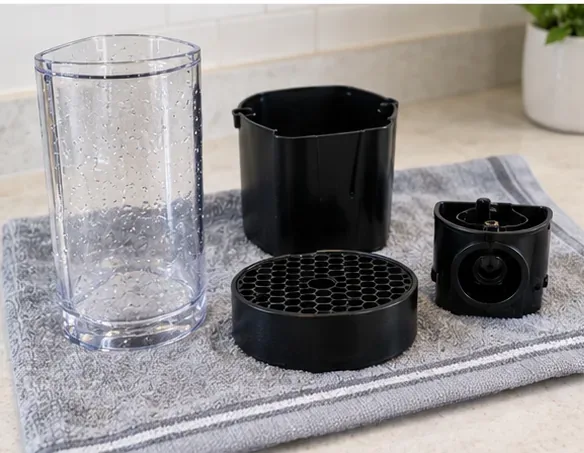 A rinsed water tank, capsule container, drip grid, and small removable coffee-machine parts drying on a clean towel.