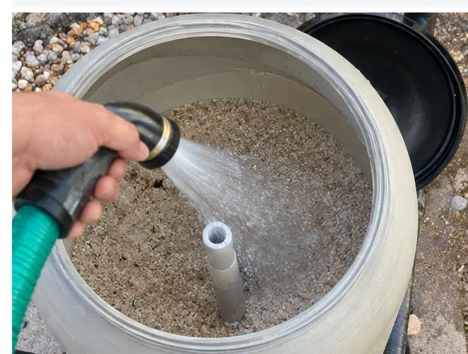 Top-down view into an open pool sand filter while a garden hose sprays across the exposed filter media around a central standpipe.