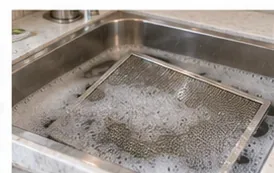 A rectangular metal range hood filter soaking flat in a sink filled with sudsy water