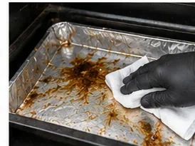 A gloved hand wiping greasy residue from a foil-covered pellet-grill drip tray