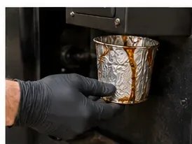 A gloved hand removing a grease-stained metal collection bucket from the side of a pellet grill
