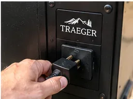 A hand unplugging the power cord from the side of a black Traeger pellet grill