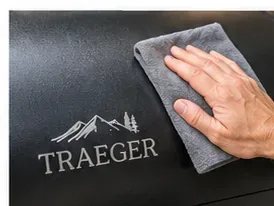 A hand wiping the black exterior lid of a Traeger pellet grill with a soft gray cloth