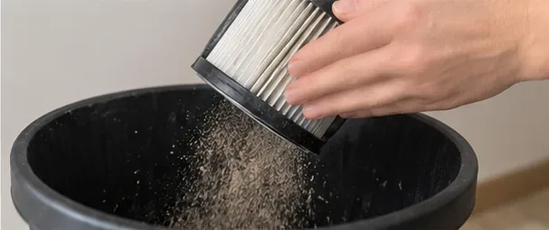 A hand lightly taps a pleated vacuum filter over a black trash can while loose dust falls away.