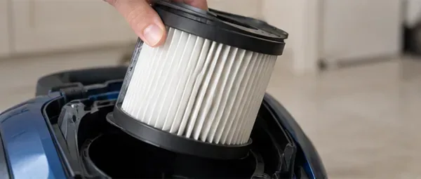 A hand lifts a white pleated cylindrical filter straight out of an open vacuum filter compartment.
