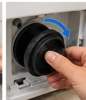 A hand turning a large round washing-machine pump filter counterclockwise inside the lower service opening