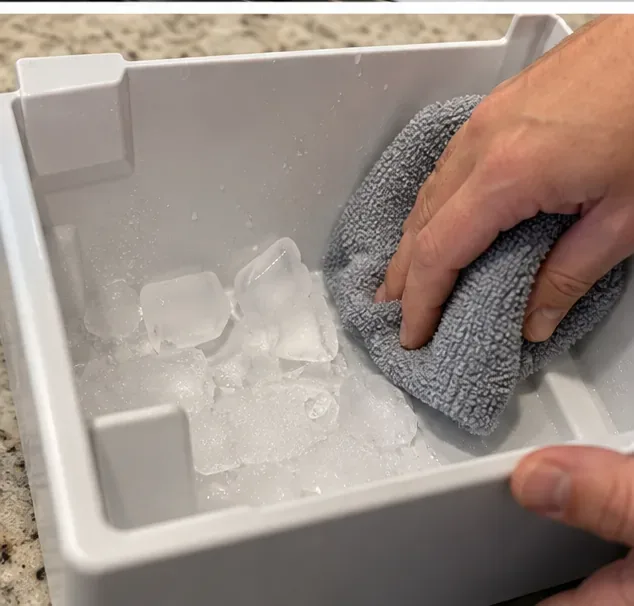 A microfiber cloth melting and wiping away ice stuck inside a removed freezer ice bin.