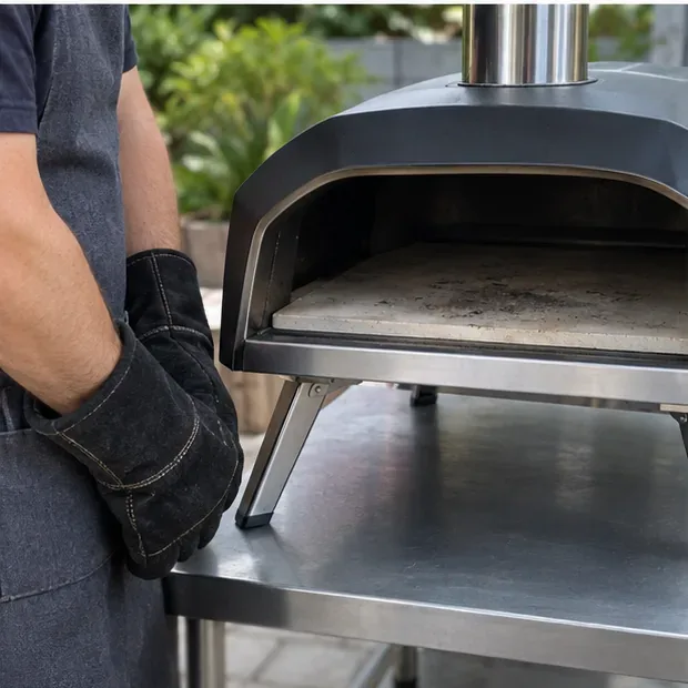 A person wearing heat-resistant gloves stands beside a cooled outdoor pizza oven with the stone visible inside.