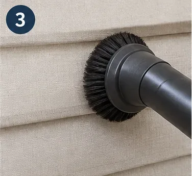 A low-suction vacuum brush gently removing dust from beige fabric blinds