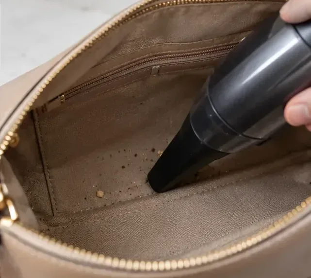 A small handheld vacuum removing crumbs from the bottom and seams of a fabric-lined purse.