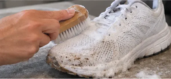 A soft brush working mild soapy water over a dirty white mesh running shoe.