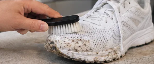 A soft-bristle brush removing dried mud from the toe of a white mesh running shoe.