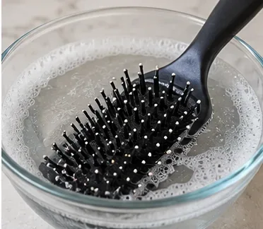 A black plastic paddle hairbrush resting in a bowl of warm soapy water