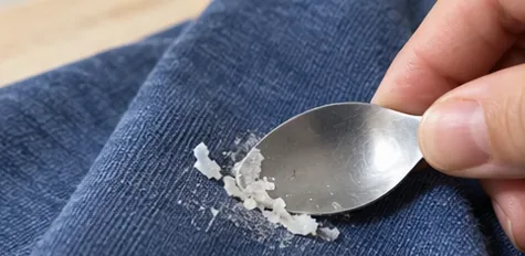 A spoon gently lifting brittle dried glue from blue fabric.