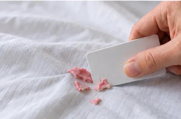 A hand using a plastic card to lift hardened chewing gum from a white bed sheet