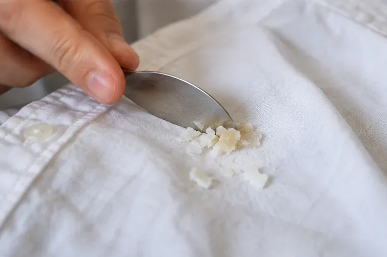 A dull metal spoon scraping brittle hardened wax from the surface of a white shirt