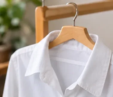 A freshly cleaned white shirt hanging on a wooden hanger to air dry indoors.