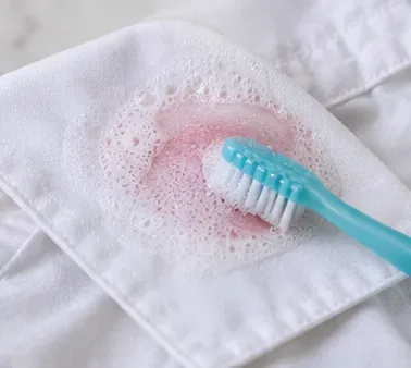 A soft toothbrush gently working suds over a fading pink lipstick mark on white fabric.