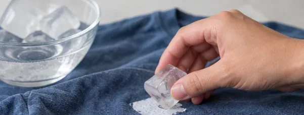 Hand holding an ice cube over thick sticker residue on a blue shirt