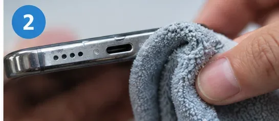 A hand wiping the outside edge of a smartphone near the charging port with a soft microfiber cloth.
