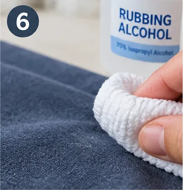 A hand dabbing blue fabric with a white cloth beside a bottle labeled 70 percent rubbing alcohol