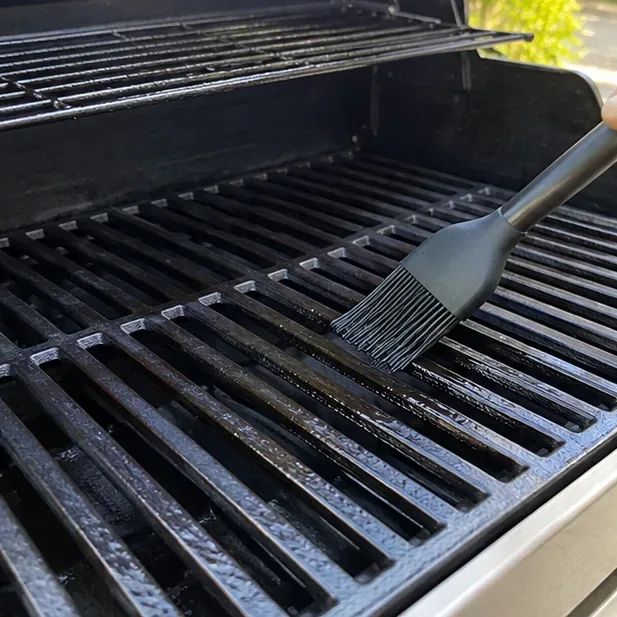 A silicone brush applying a second thin coat of oil across cast-iron grates inside a gas grill.