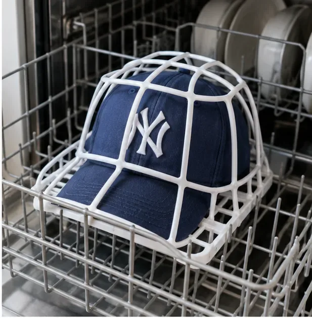 A navy baseball cap secured in a white cap frame on a dishwasher rack with dishes visible behind it