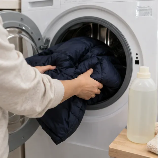 A dark navy puffer jacket being loaded by hand into a front-loading washing machine beside a bottle of gentle detergent.