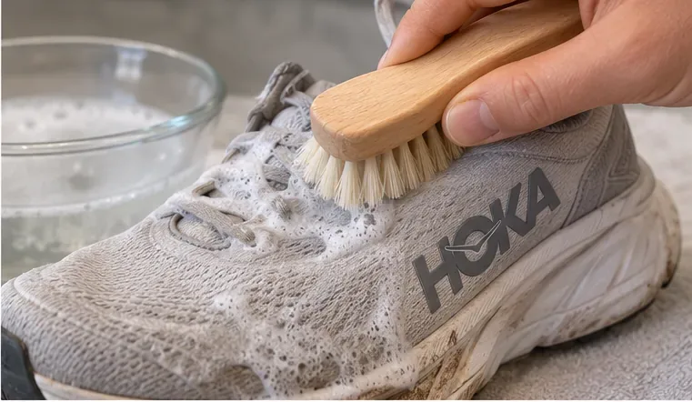 A soft-bristle brush gently working mild soap suds into the mesh upper of a light-colored HOKA running shoe