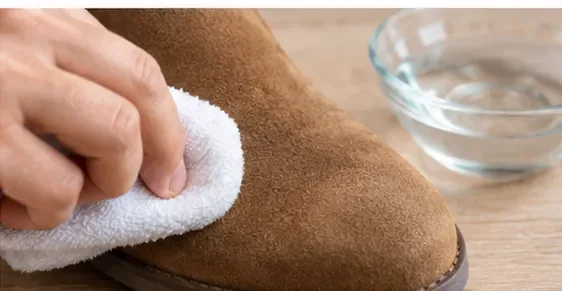 A white cloth lightly spot-cleaning the toe of a brown suede boot beside a small bowl of water
