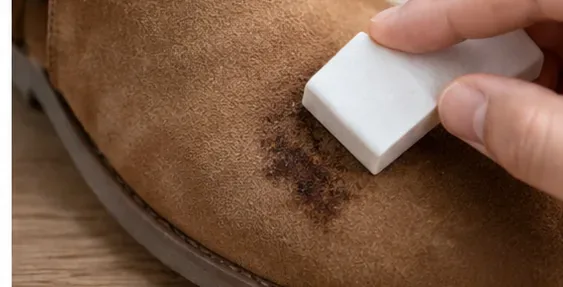 A white suede eraser being rubbed gently over a dark scuff on a brown suede boot