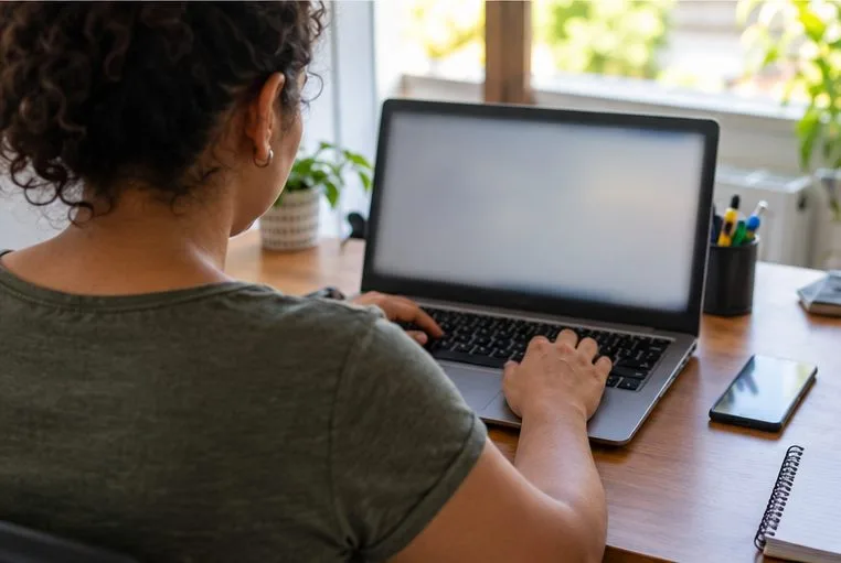 Uma microempreendedora preenche dados no notebook, com a tela fora de foco e sem informações legíveis.