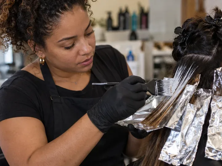Colorista aplica tonalizante com pincel em uma mecha separada, usando luvas e papel de proteção