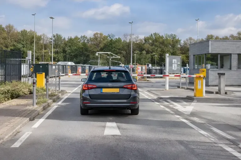 Een auto nadert een toegangspoort van een gemeentelijke milieustraat met slagboom en rijstroken