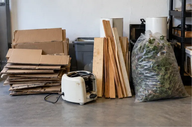 Gesorteerd afval in een garage: plat karton, losse houten planken, een klein elektrisch apparaat en een zak tuinafval staan apart klaar.