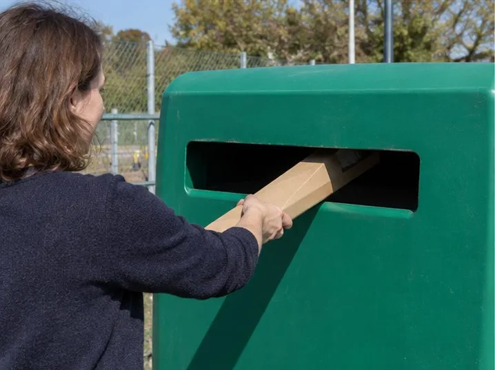 Une personne dépose un long emballage en carton dans l’ouverture d’un conteneur de collecte extérieur