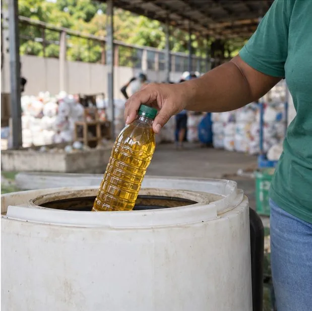 Uma pessoa leva uma garrafa PET tampada com óleo usado até um recipiente de coleta em um espaço comunitário.