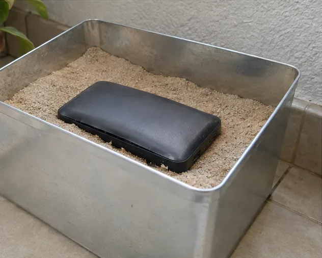 Power bank estufado apoiado em areia dentro de uma bandeja metálica aberta e resistente, usado apenas como isolamento temporário