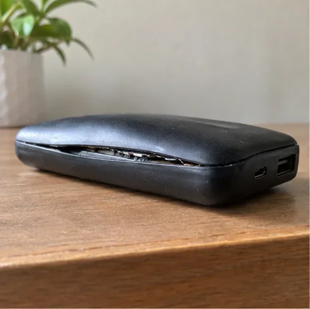 Power bank preto com a carcaça arqueada e uma abertura na junção, apoiado sozinho sobre uma mesa de madeira