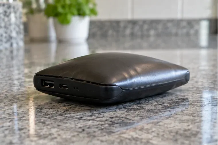 Power bank preto visivelmente estufado, com a carcaça abaulada, apoiado sobre uma bancada de pedra