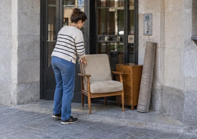 Una vecina coloca de forma ordenada un sillón, una cajonera y una alfombra junto al portal del edificio.