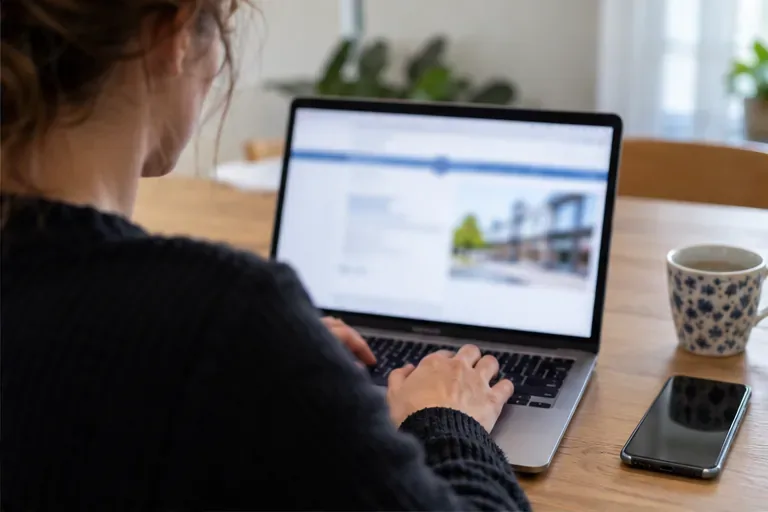 Een vrouw gebruikt thuis een laptop met een onscherp scherm; een telefoon ligt naast haar.