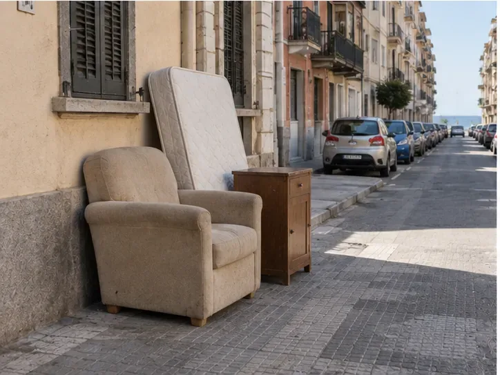 Poltrona, materasso e piccolo mobile disposti ordinatamente vicino a un edificio senza occupare la carreggiata