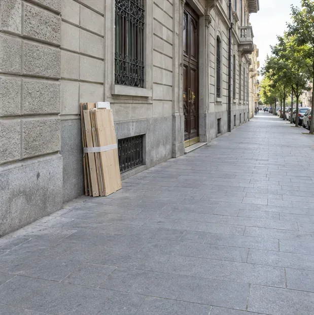 Pannelli di legno legati insieme sono appoggiati vicino alla facciata, sul bordo di un marciapiede ampio e libero.