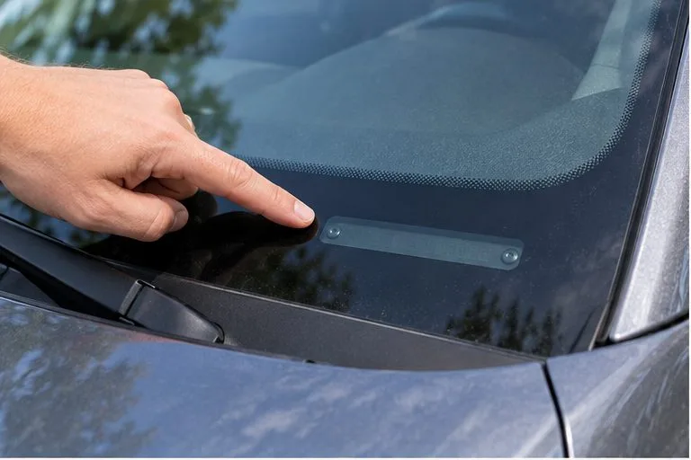 A hand points to the VIN plate at the lower edge of a car windshield; the characters are not legible.