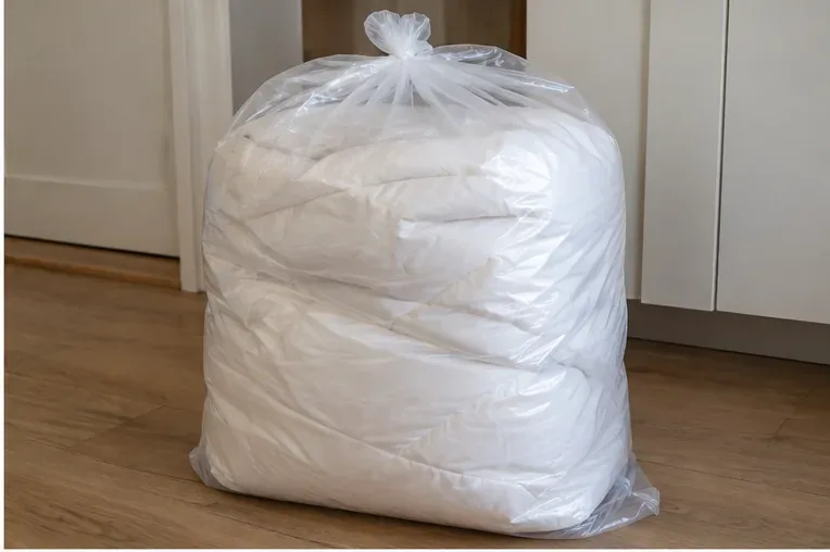 Een opgevouwen wit dekbed zit volledig in een doorzichtige, dichtgebonden plastic zak.