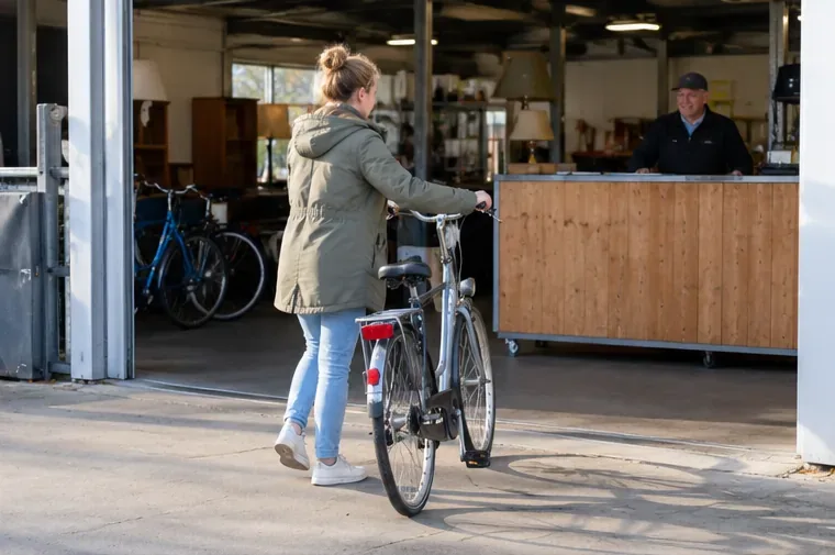 Een vrouw brengt een nette stadsfiets naar een overdekte innamebalie waar een medewerker klaarstaat.