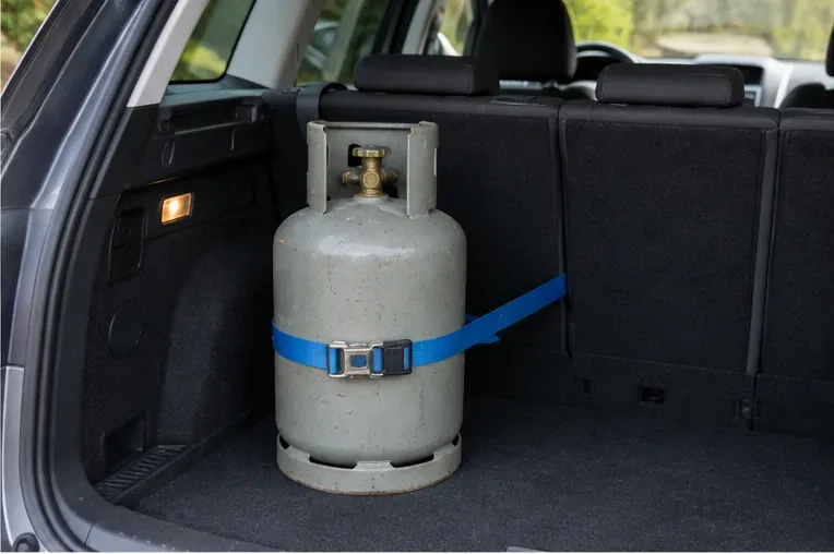 Een grijze gasfles staat rechtop in de bagageruimte van een auto en is met een stevige blauwe spanband tegen verschuiven vastgezet.
